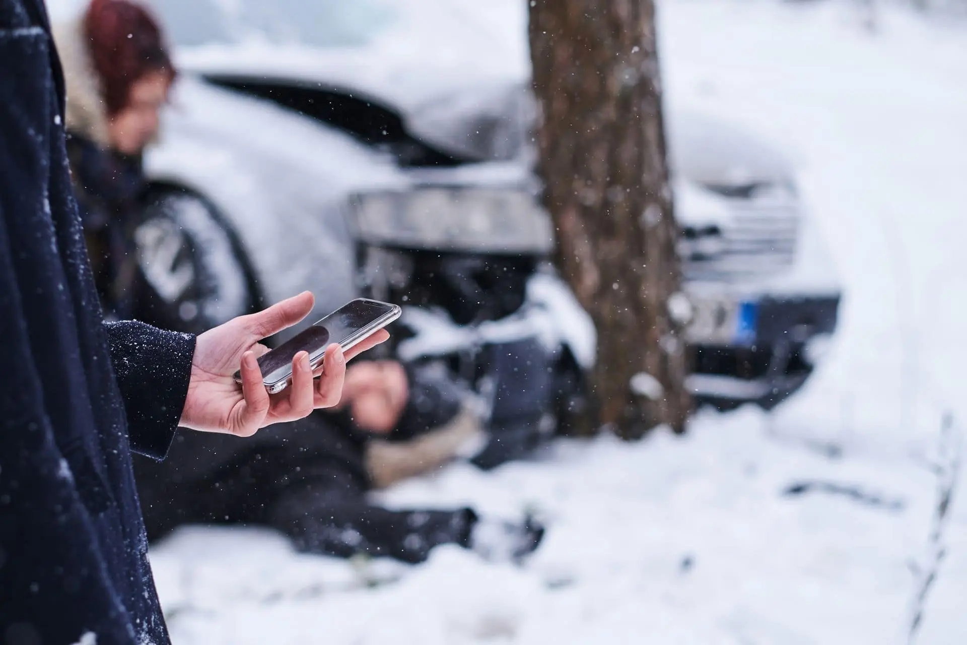 Persona con el celular en la mano junto a un auto chocado contra un árbol en la nieve