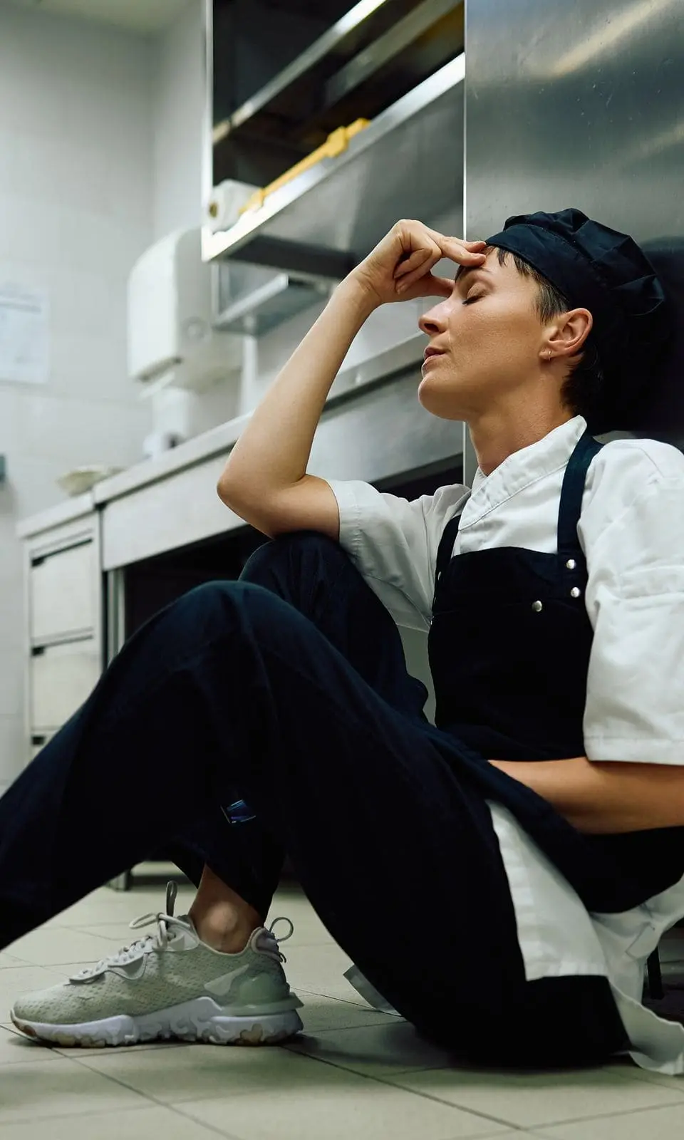 Trabajadora de cocina sentada en el piso con la mano en la frente después de un accidente laboral